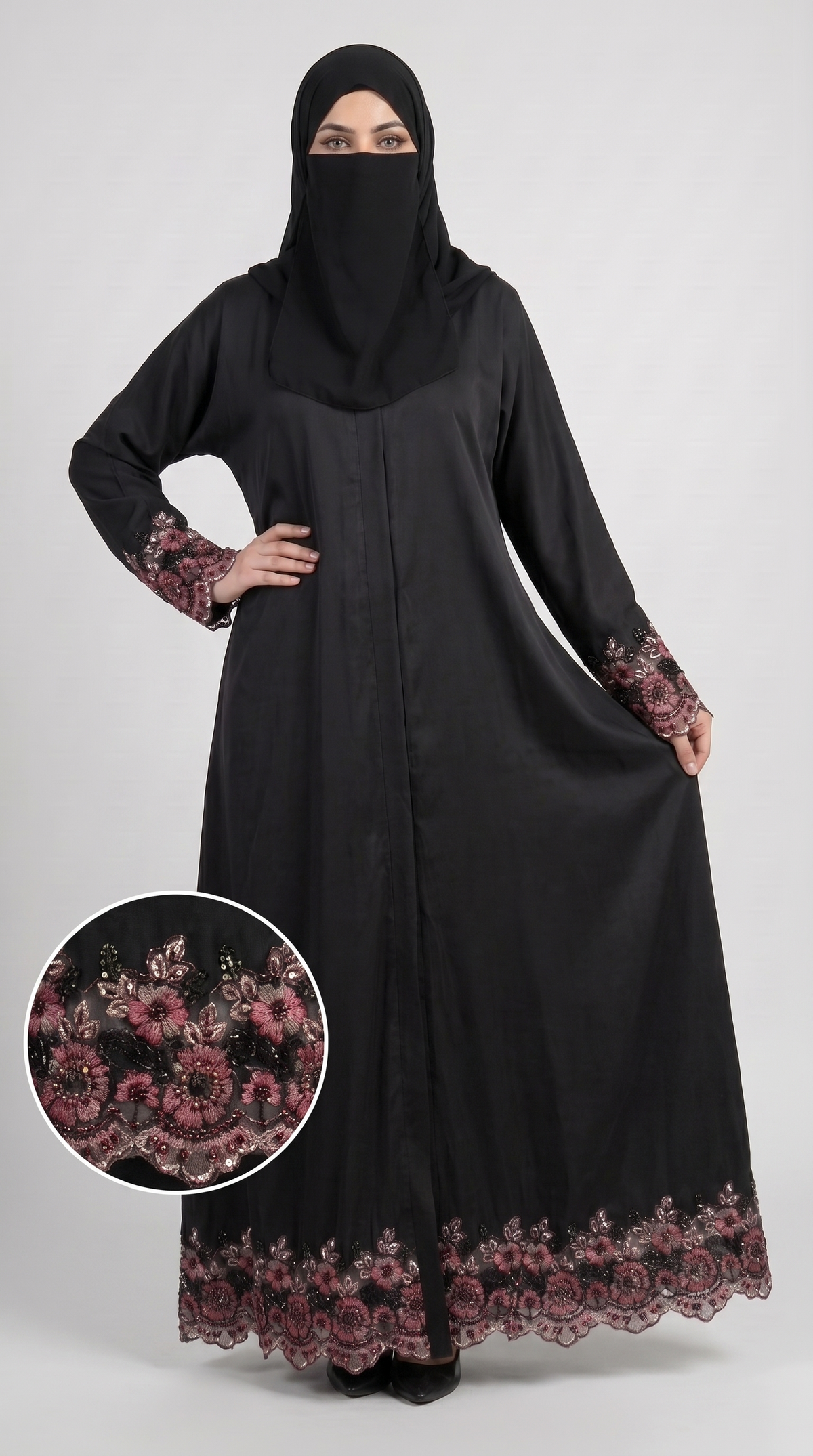 A woman modeling a black long abaya with pink floral lace trim on the sleeves and hem, paired with a matching black hijab, against a light gray studio background. A circular inset shows a close-up of the pink lace pattern.