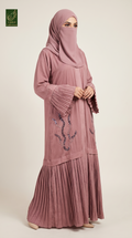 A woman modeling a dusty rose pink modest outfit consisting of a long-sleeved abaya with decorative sequined embroidery on the front and flared cuffs, paired with a matching hijab. The image is shot against a plain light beige studio background.