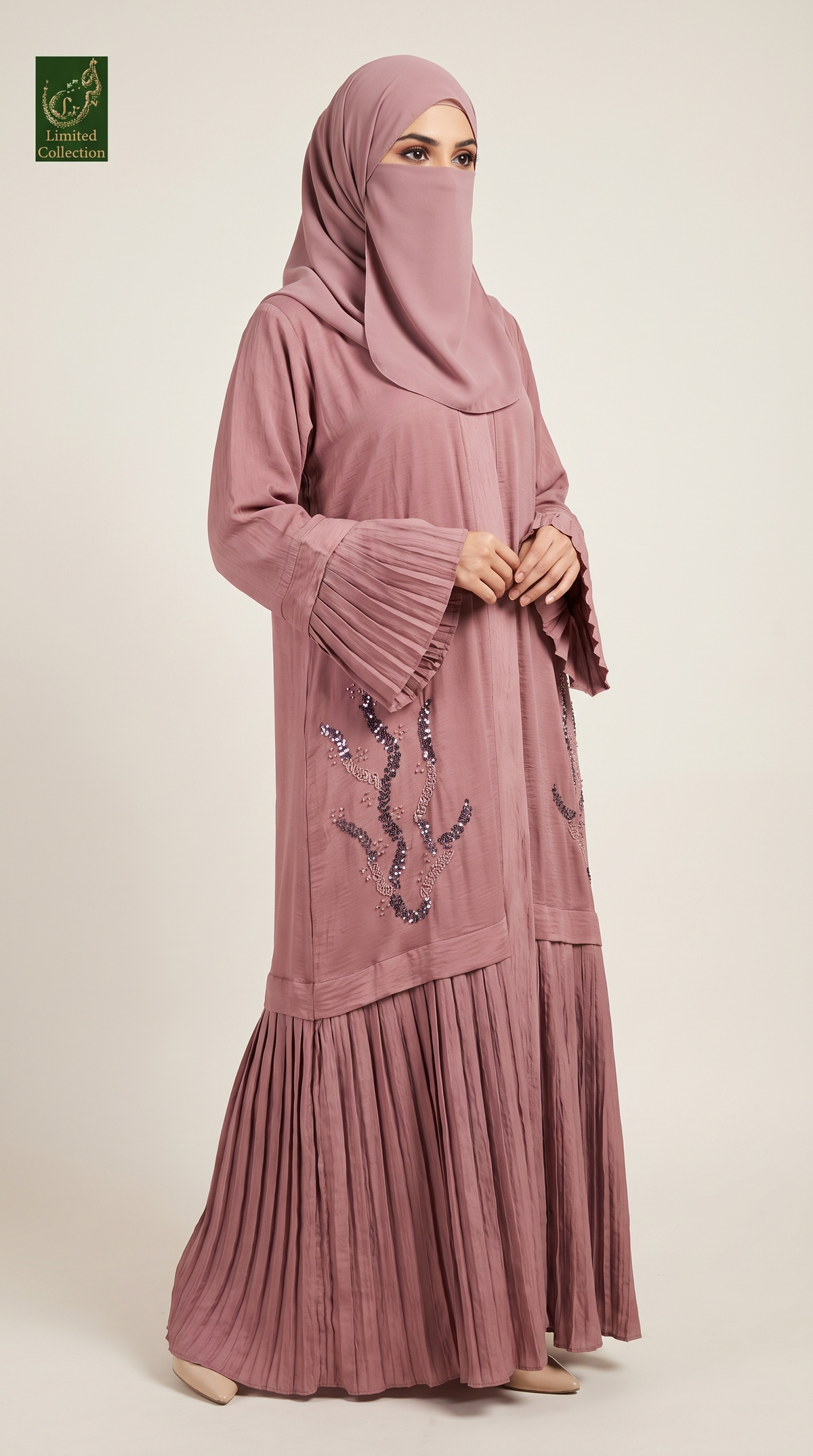 A woman modeling a dusty rose pink modest outfit consisting of a long-sleeved abaya with decorative sequined embroidery on the front and flared cuffs, paired with a matching hijab. The image is shot against a plain light beige studio background.