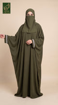 A woman wearing a flowing olive green abaya with a matching hijab and a small silver mirror embellishment on the sleeve, standing against a solid beige background. A logo for 'Limited Collection' is visible in the top-left corner.