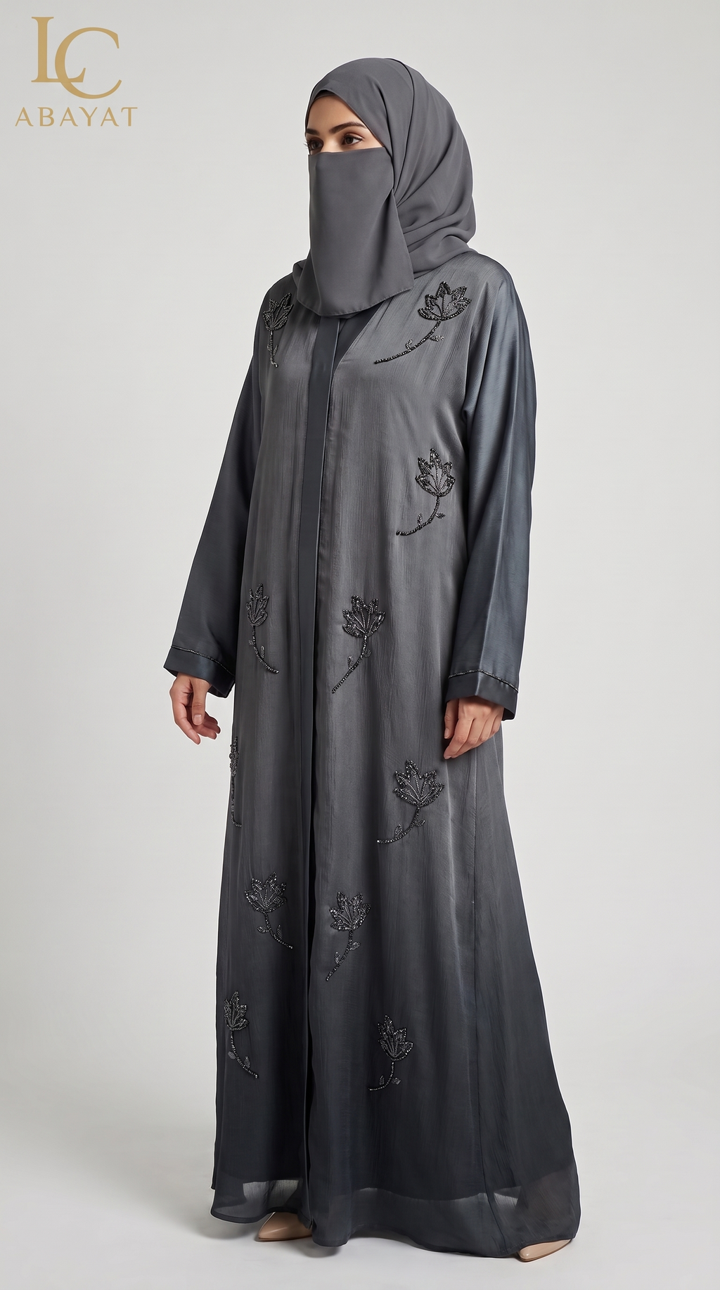 A woman wearing a dark gray abaya with black floral embroidery and a matching gray hijab, standing against a light gray studio background. The LC Abayat brand logo is visible in the top left corner.