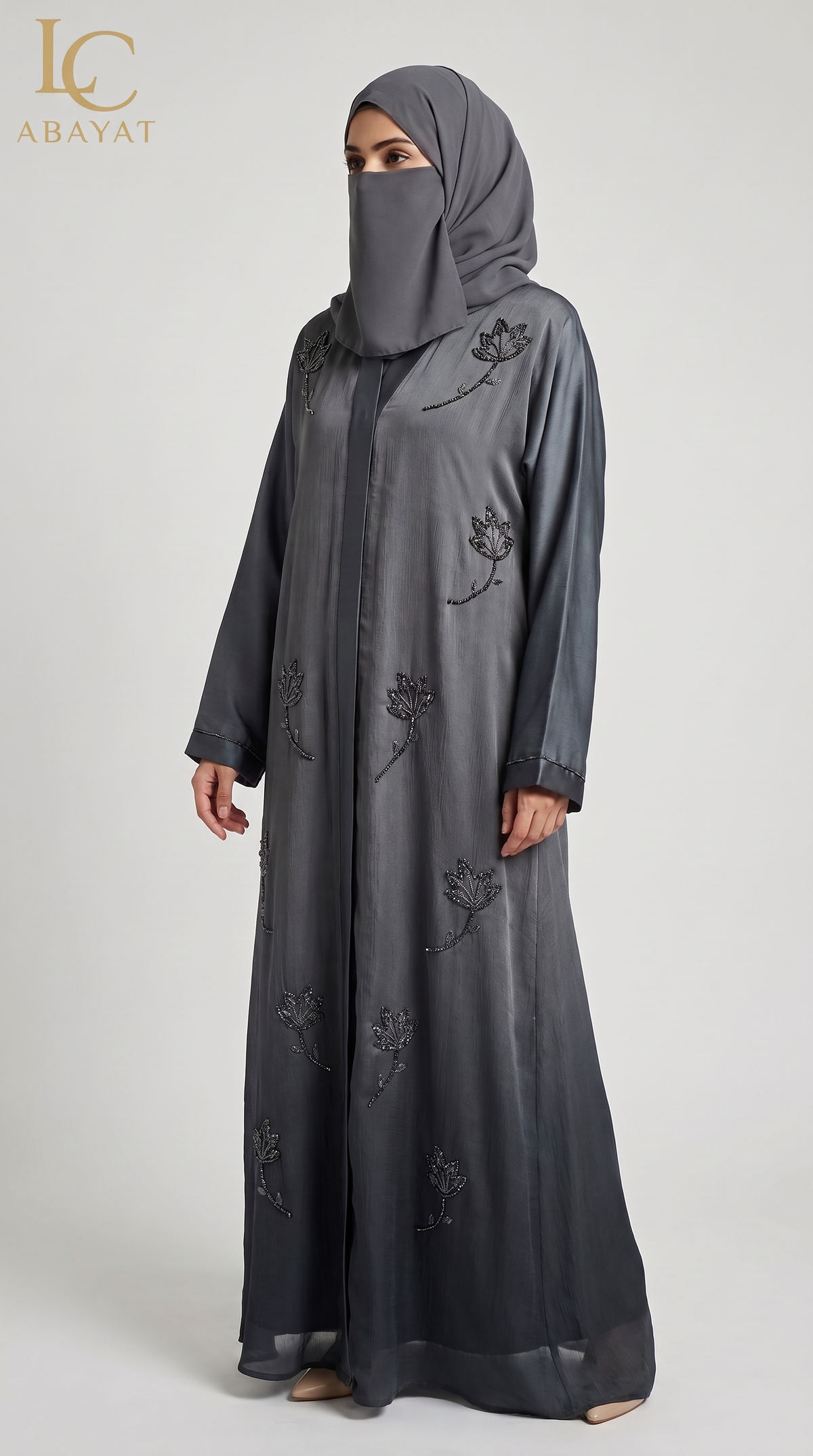 A woman wearing a dark gray abaya with black floral embroidery and a matching gray hijab, standing against a light gray studio background. The LC Abayat brand logo is visible in the top left corner.