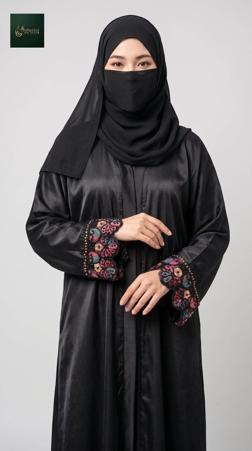 A woman wearing a black nida abaya with colorful floral cutdana beaded handwork on the cuffs and a matching black hijab, photographed against a light gray studio background.