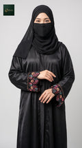 A woman wearing a black nida abaya with colorful floral cutdana beaded handwork on the cuffs and a matching black hijab, photographed against a light gray studio background.