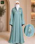 A woman wearing a long, flowing powder blue abaya with white jasmine embroidery on the chest and sleeves, paired with a white hijab. The image features a circular inset showing a close-up of the embroidery detail and a logo in the top left corner.