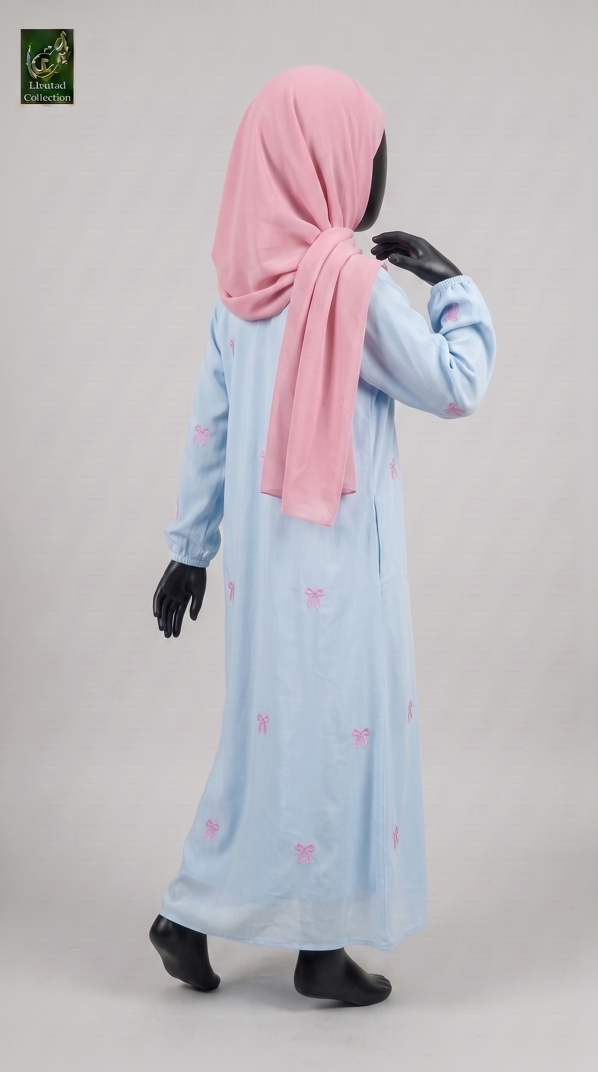 A child model wearing a light blue long-sleeve abaya dress with small pink bow prints and a pink hijab, shown from the back against a light gray studio background. Black mannequin feet and hands are visible.