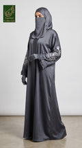 A woman modeling a dark gray satin abaya with silver-gray embroidered sleeves, a matching gray hijab, and gray gloves, standing against a plain light gray wall.