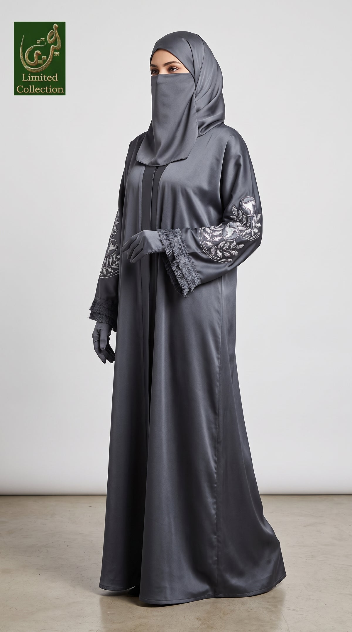 A woman modeling a dark gray satin abaya with silver-gray embroidered sleeves, a matching gray hijab, and gray gloves, standing against a plain light gray wall.