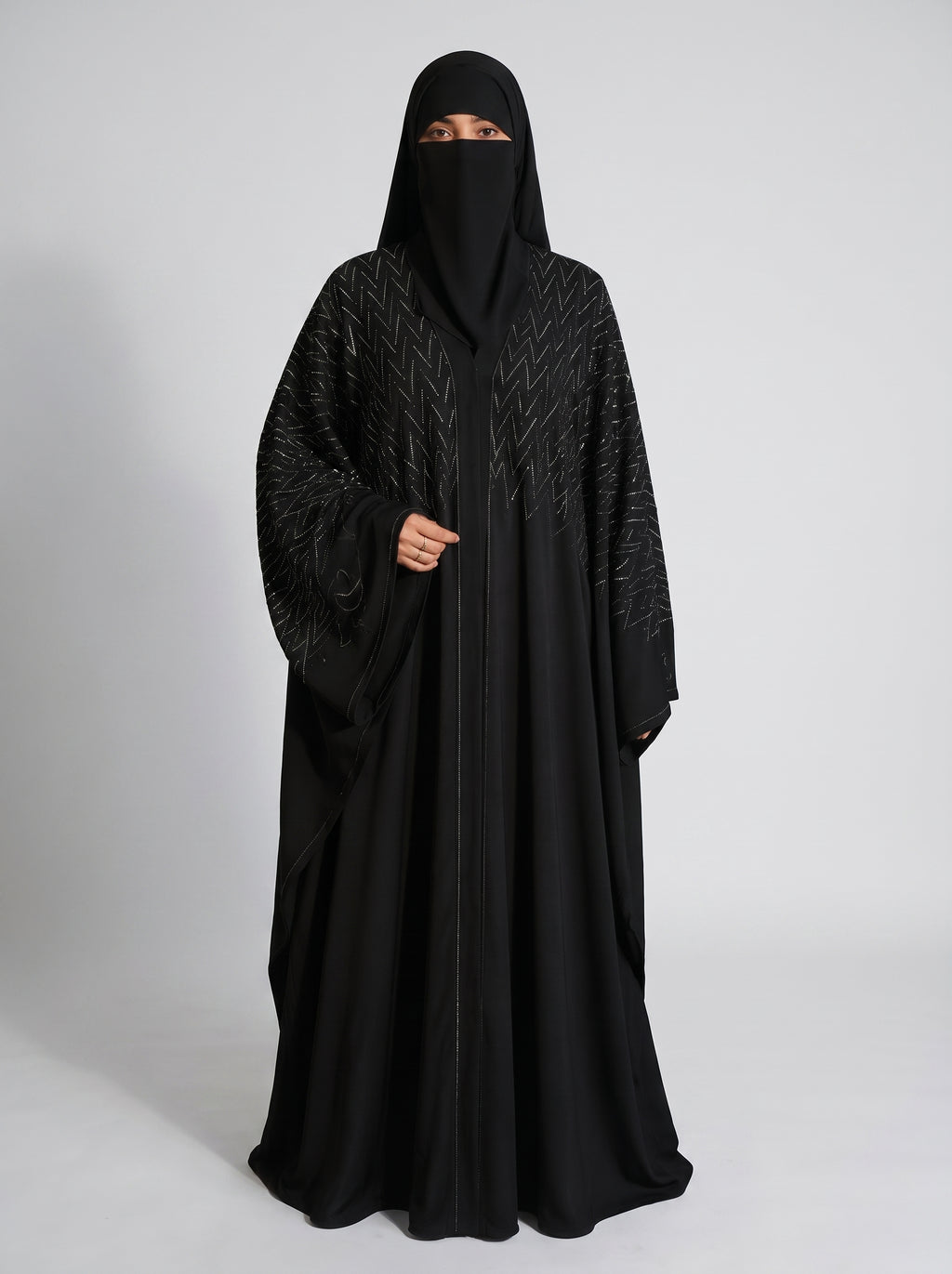 A woman wearing a full black abaya and matching face covering, standing against a light gray studio background. The abaya has subtle tonal geometric embroidery or textured patterning on the upper chest and sleeves.