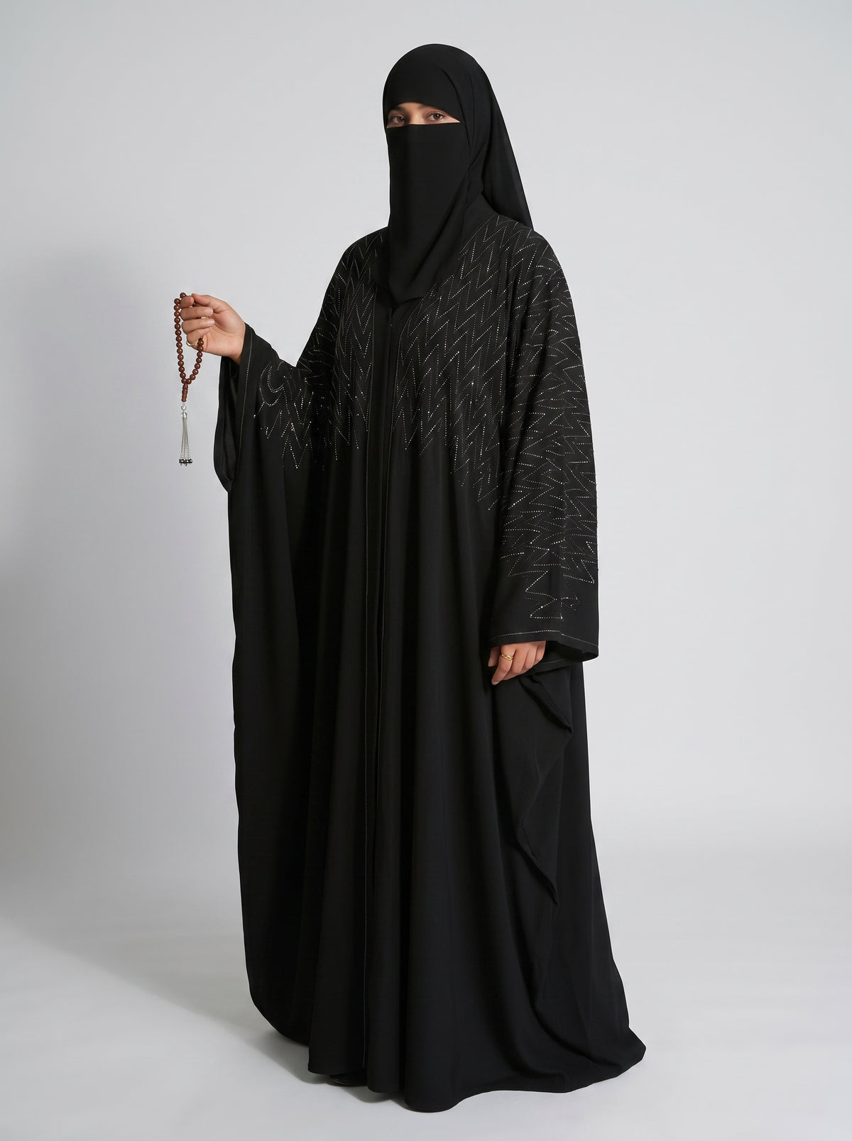 A woman wearing a full black abaya and matching face covering, holding a strand of prayer beads, photographed against a light gray studio background.