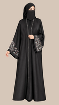A woman wearing a black abaya with silver floral embroidery on the wide sleeves and a matching black hijab, standing against a solid beige studio background. The LC ABAYAT logo is visible in the top left corner.