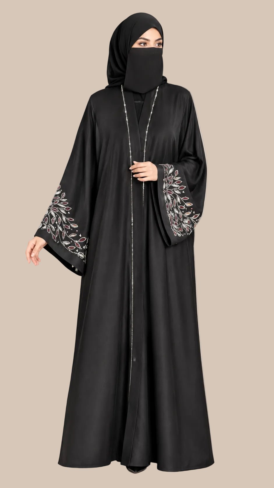 A woman wearing a black abaya with silver floral embroidery on the wide sleeves and a matching black hijab, standing against a solid beige studio background. The LC ABAYAT logo is visible in the top left corner.