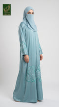 A woman modeling a light blue modest abaya with cutdana handwork consisting of a long abaya with teal floral embroidery and a matching hijab, shown against a plain light gray studio background. A 'Lamored Collection' logo is visible in the top-left corner.